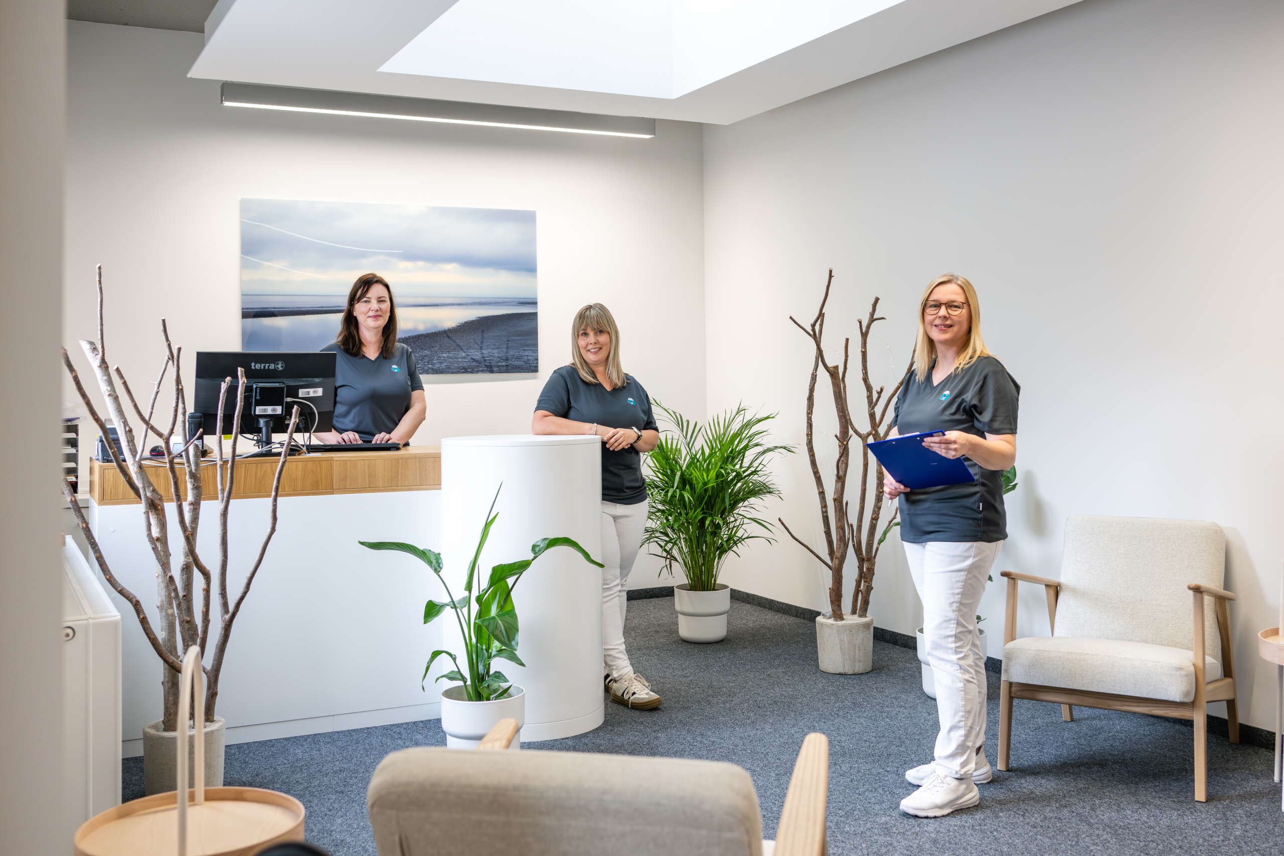 Drei Frauen in grauen T-Shirts stehen in einem Empfangsbereich mit Pflanzen und einem Empfangstresen.