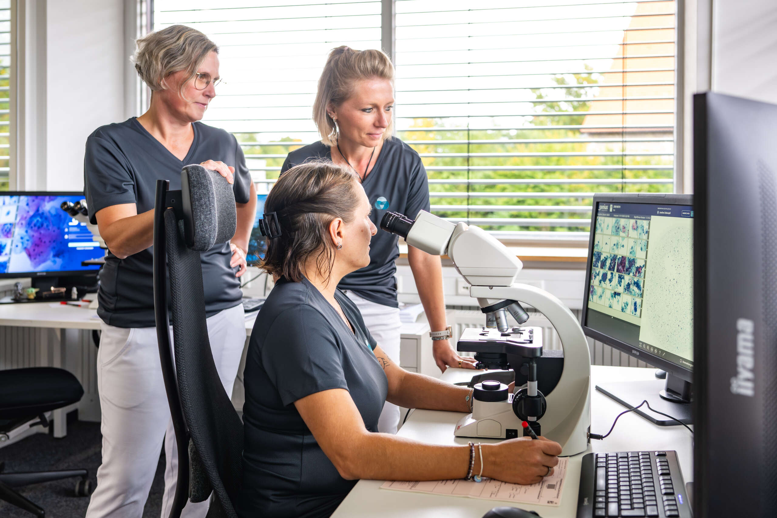 Drei Mitarbeiterinnen in einem Labor, eine schaut durch ein Mikroskop, während die anderen beobachten.