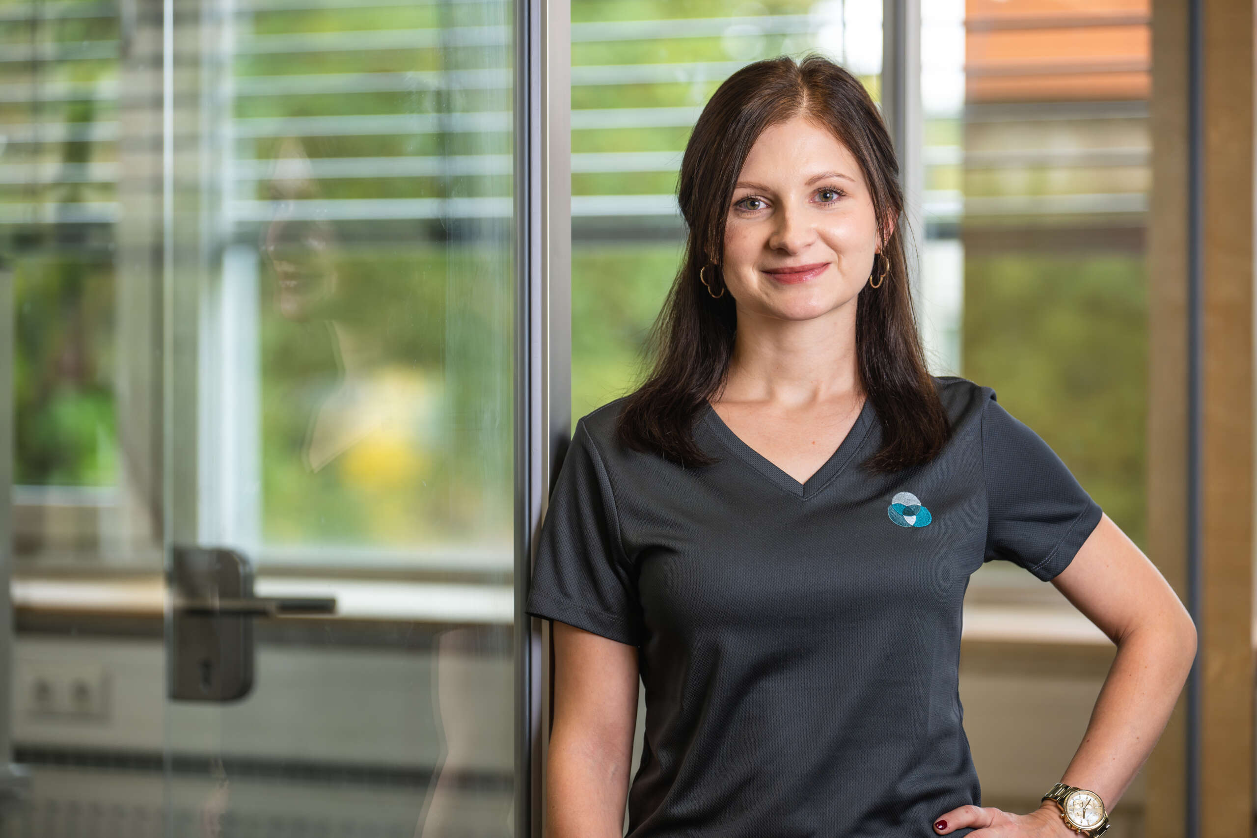 Frau mit langen, dunklen Haaren trägt ein schwarzes T-Shirt mit einem Logo und steht vor einer Glaswand.