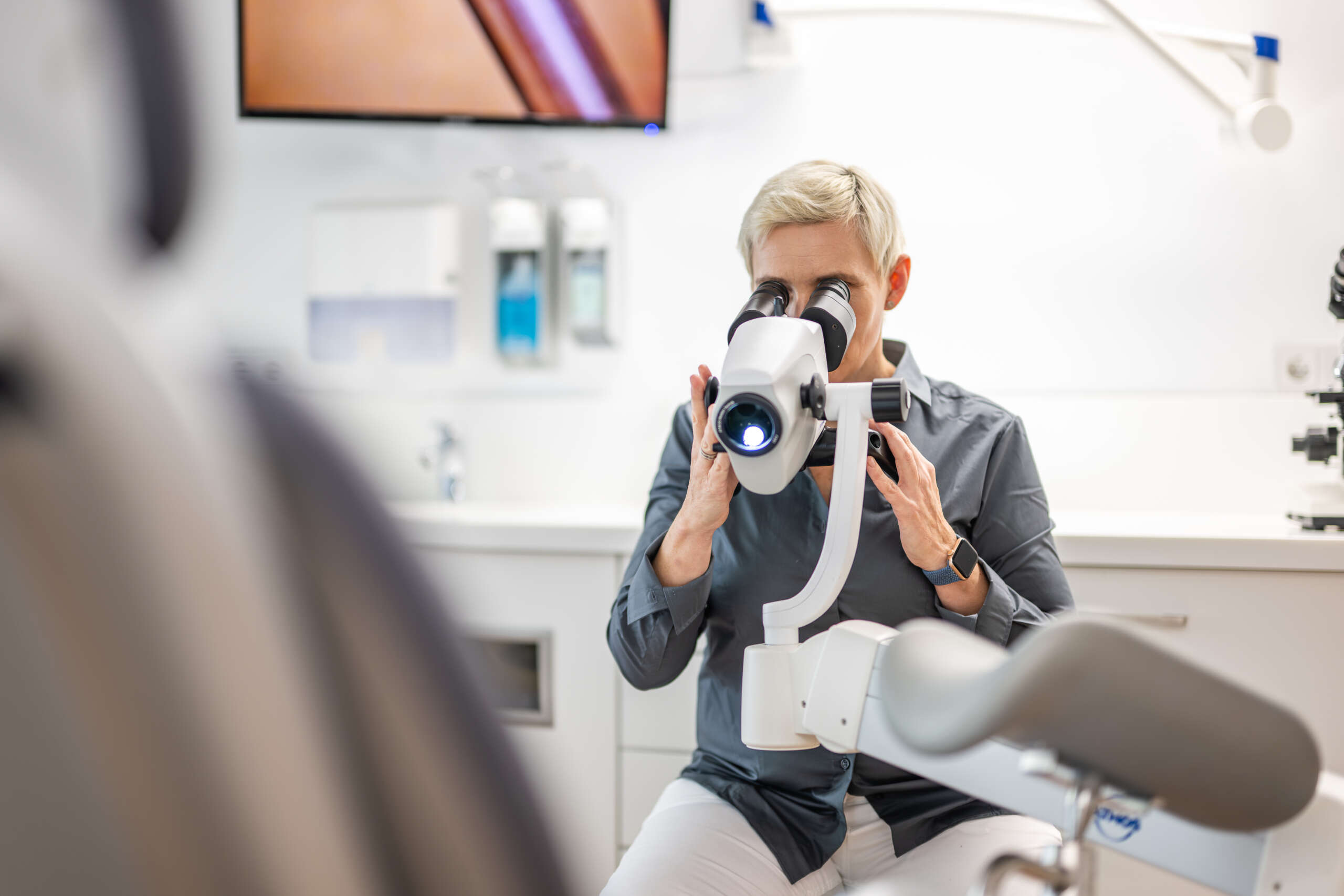 Eine Person, die ein Mikroskop in einer medizinischen Einrichtung benutzt, mit einem Bildschirm im Hintergrund.