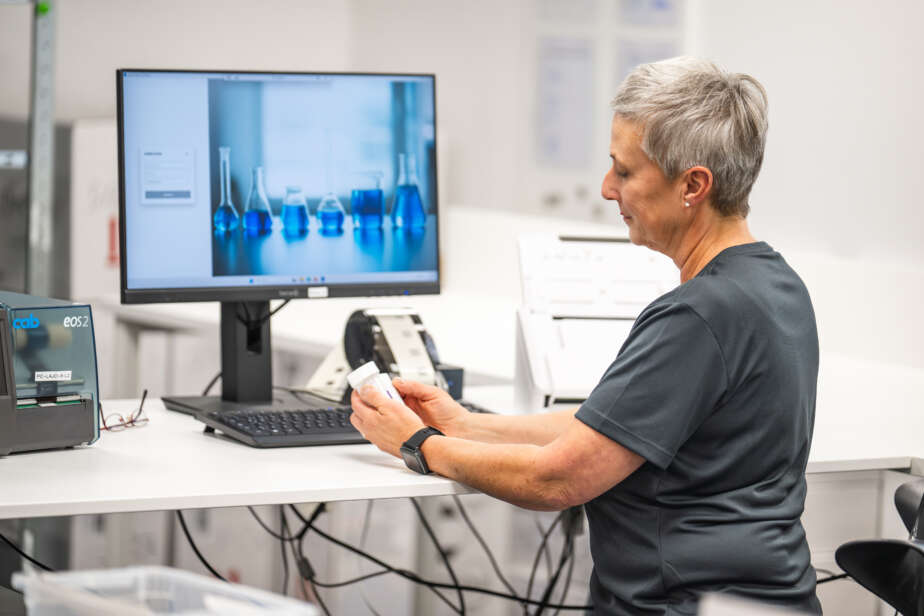 Laborantin untersucht eine Probe vor einem Computer mit blauen Flüssigkeiten in Glasbehältern im Hintergrund.