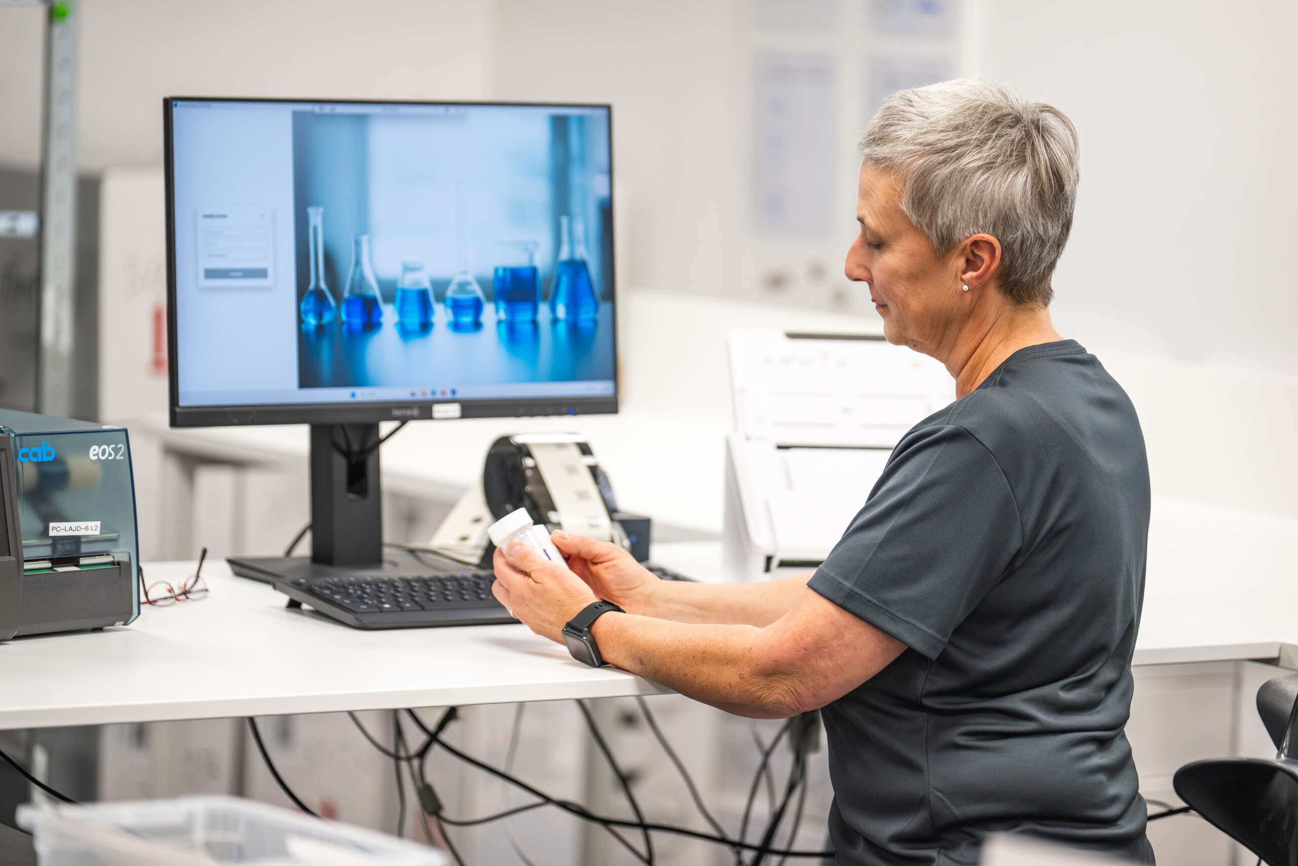 Laborantin untersucht eine Probe vor einem Computer mit blauen Flüssigkeiten in Glasbehältern im Hintergrund.