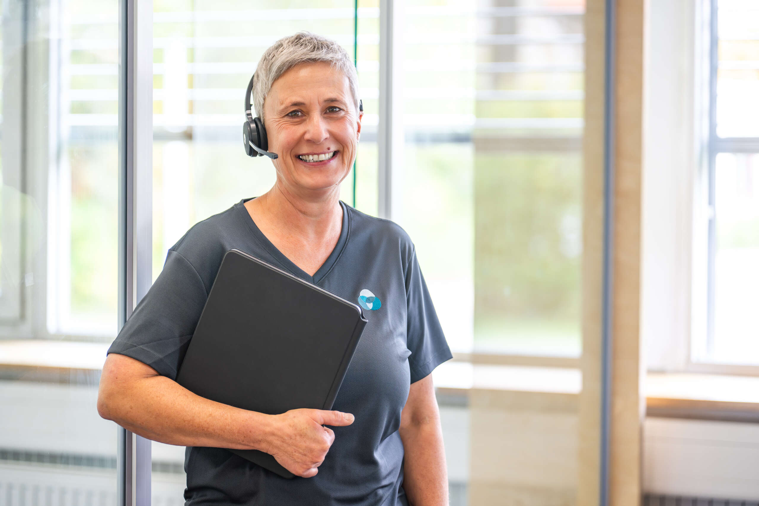 Frau mit grauem T-Shirt, Headset und schwarzem Ordner, lächelt in einem modernen Büro.