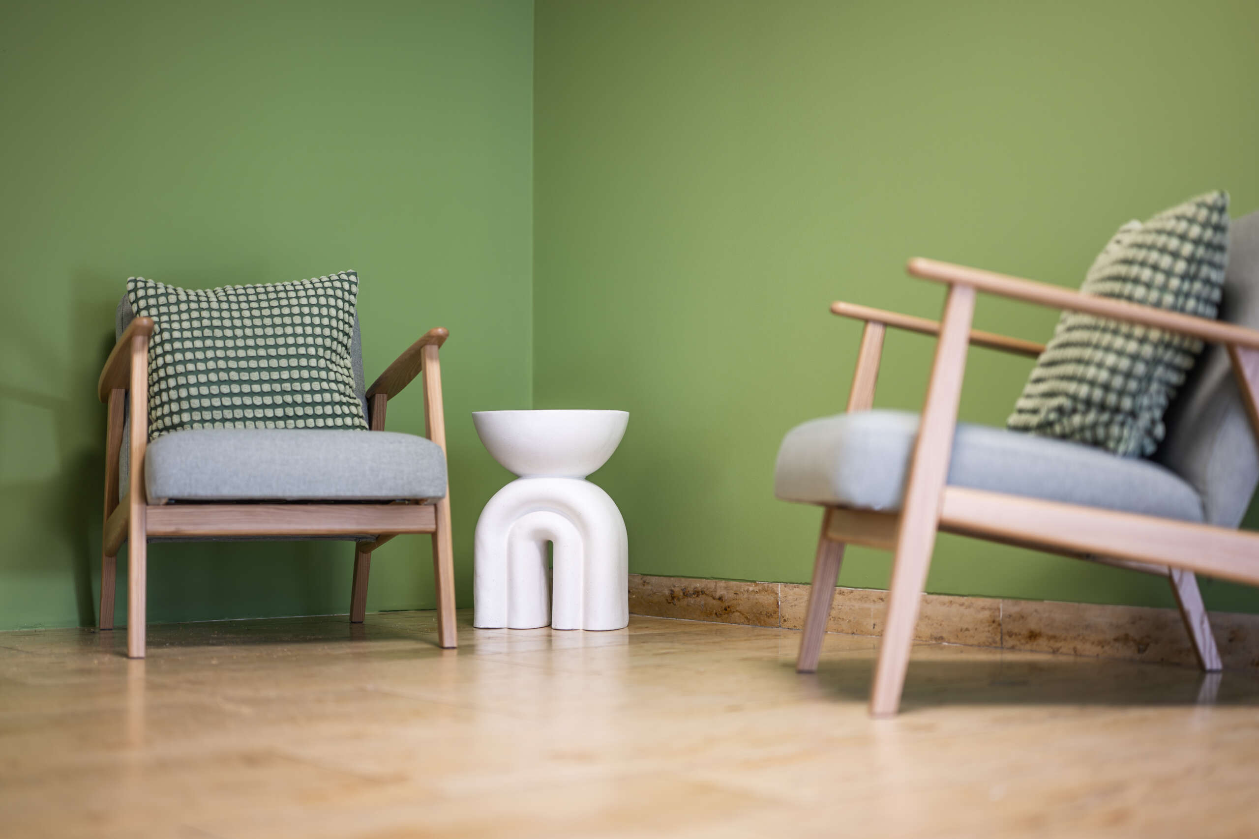 Zwei moderne Holzstühle mit grauen Kissen und einem weißen Beistelltisch in einem Raum mit grüner Wandfarbe.