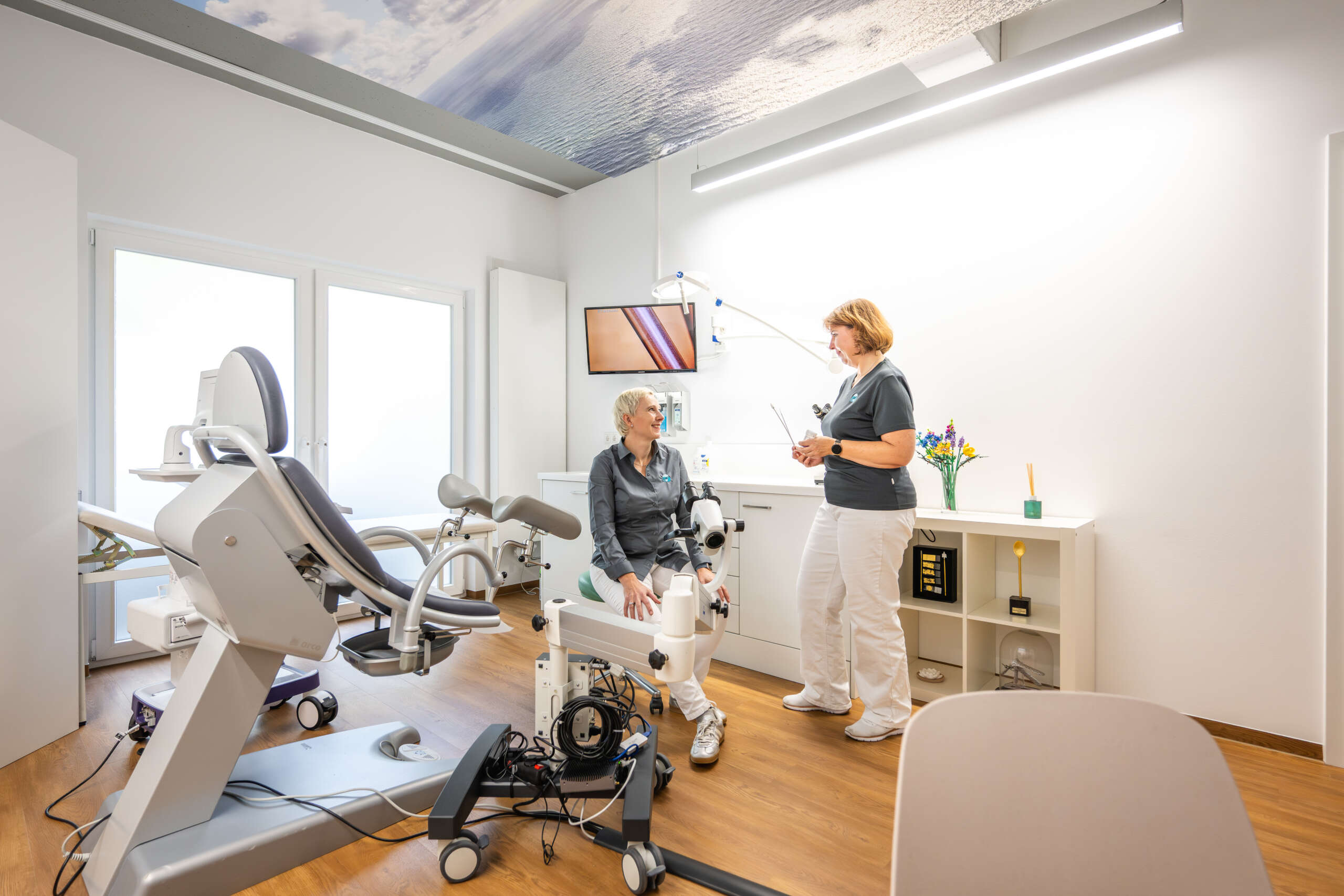 Zwei Frauen in einem modernen Behandlungsraum, eine sitzt auf einem Untersuchungstisch.