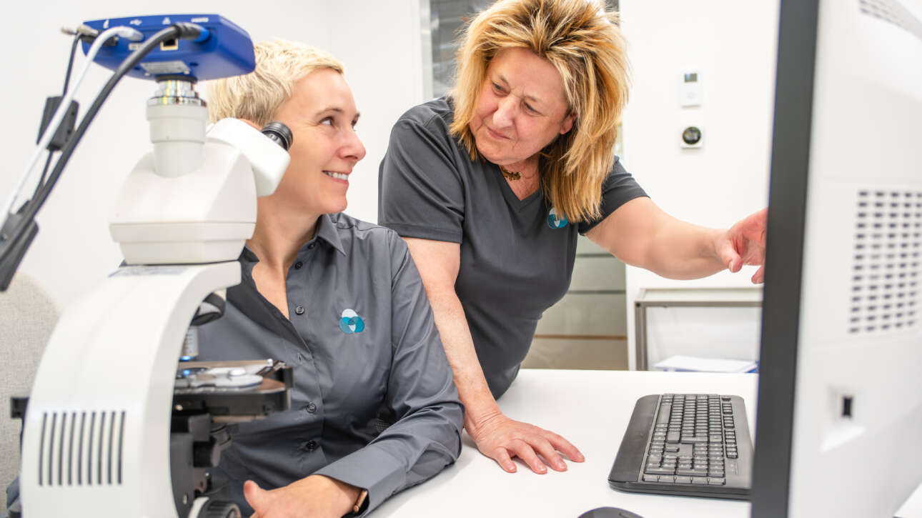 Zwei Frauen im Labor, eine schaut auf den Computerbildschirm, die andere steht neben einem Mikroskop.