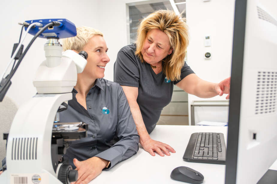 Zwei Frauen im Labor, eine schaut auf den Computerbildschirm, die andere steht neben einem Mikroskop.