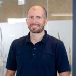 Mann mit kurzem Bart und Polohemd steht in einem modernen Büro mit Glaswänden.