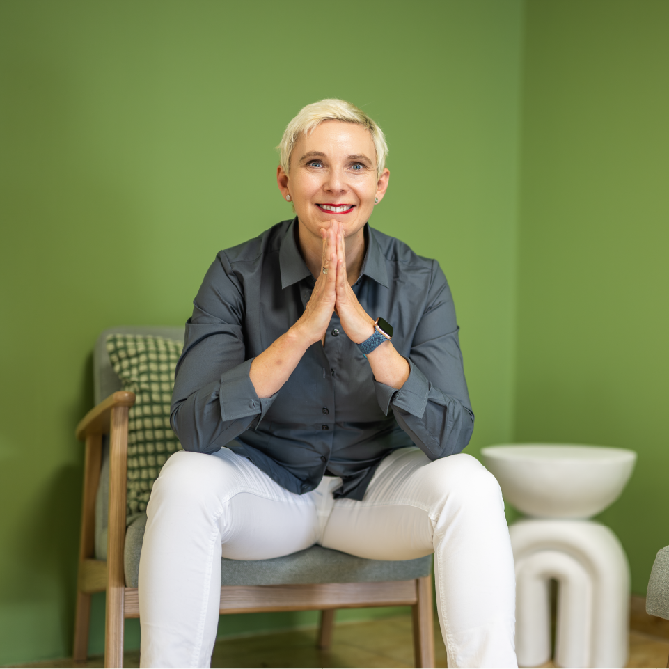 Frau mit kurzen blonden Haaren, die in einem grauen Hemd sitzt und die Hände in einer betenden Position hält.