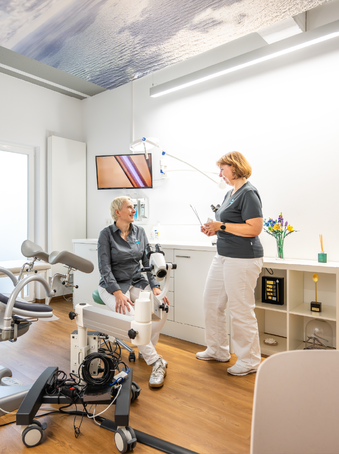 Zwei medizinische Fachkräfte in einem Behandlungsraum, eine sitzt auf einem Stuhl und die andere steht mit einem Instrument.